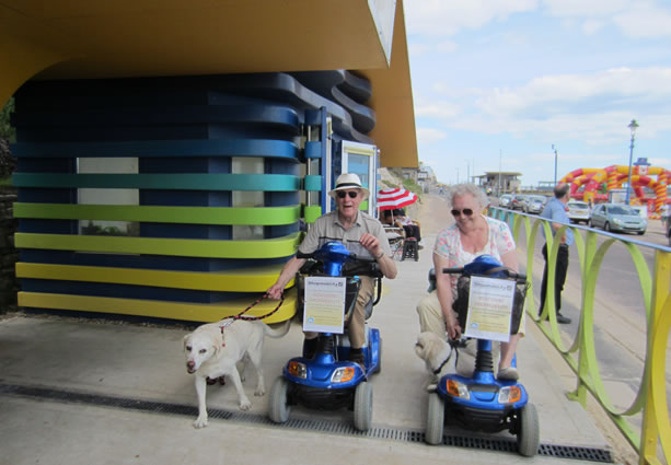 Two people on mobility scooters with a dog travelling along the seafront 